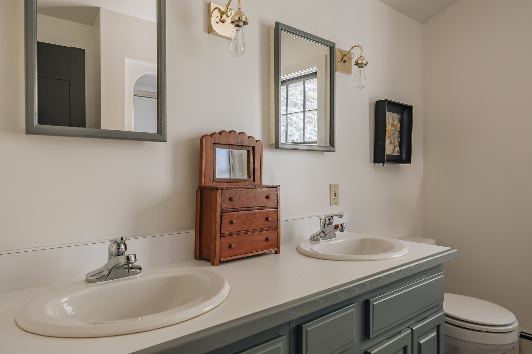 A bright bathroom featuring a double vanity with white sinks set into a light countertop, dark gray cabinets, and two mirrors with brass sconces above. A small wooden dresser with a mirror sits between the sinks.