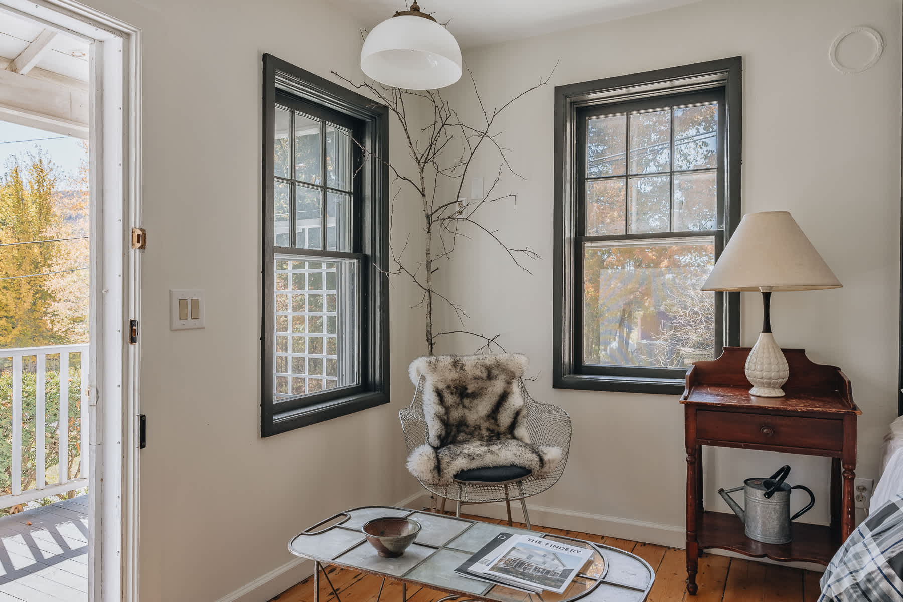 A bright corner of a room features two windows with dark trim and a door leading to an outdoor balcony. A modern wire chair with a fur throw sits next to a vintage wooden nightstand, with a decorative branch extending upwards.