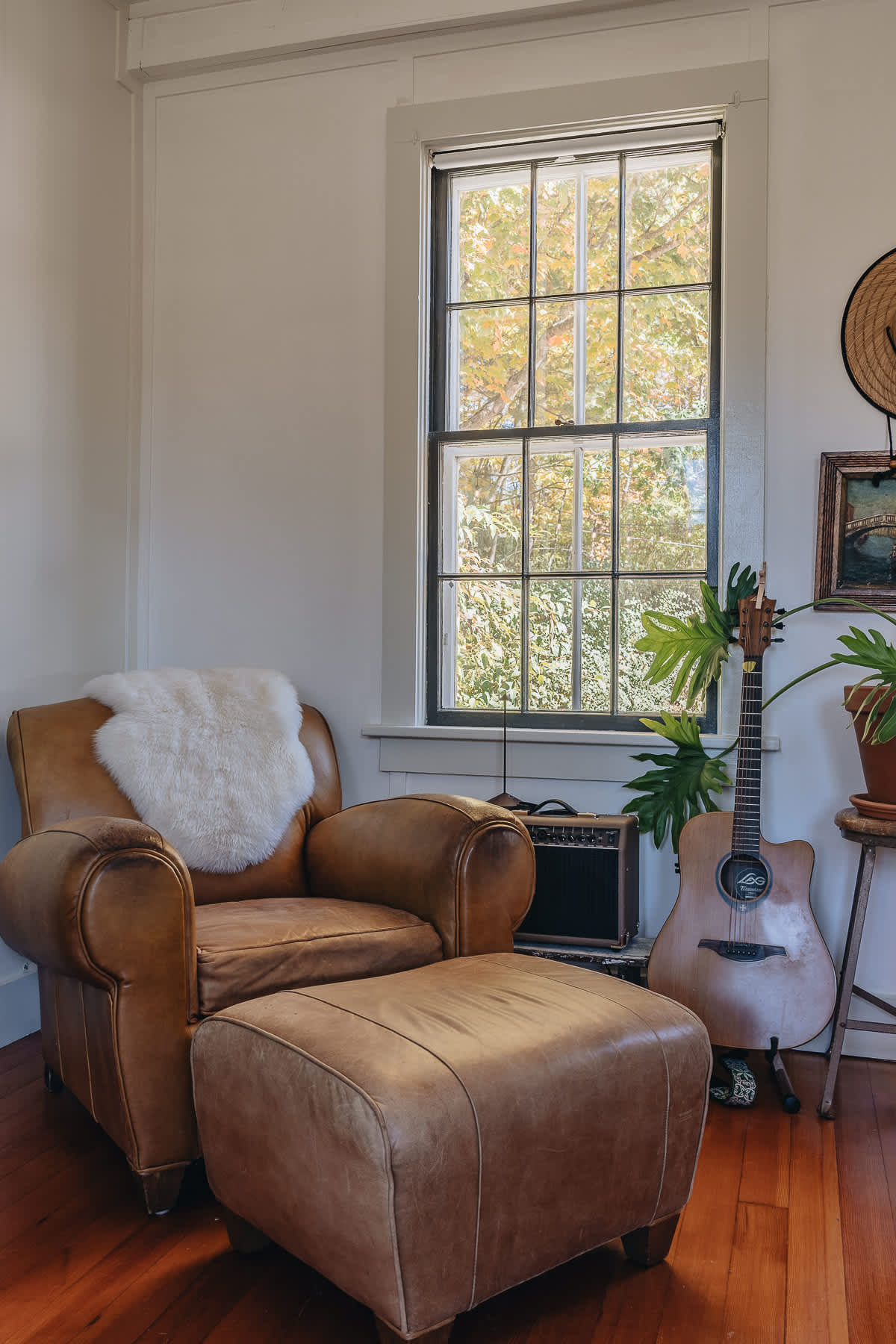 Inviting corner of a bedroom with a tan leather armchair and ottoman, a large window, and a guitar next to a potted plant.