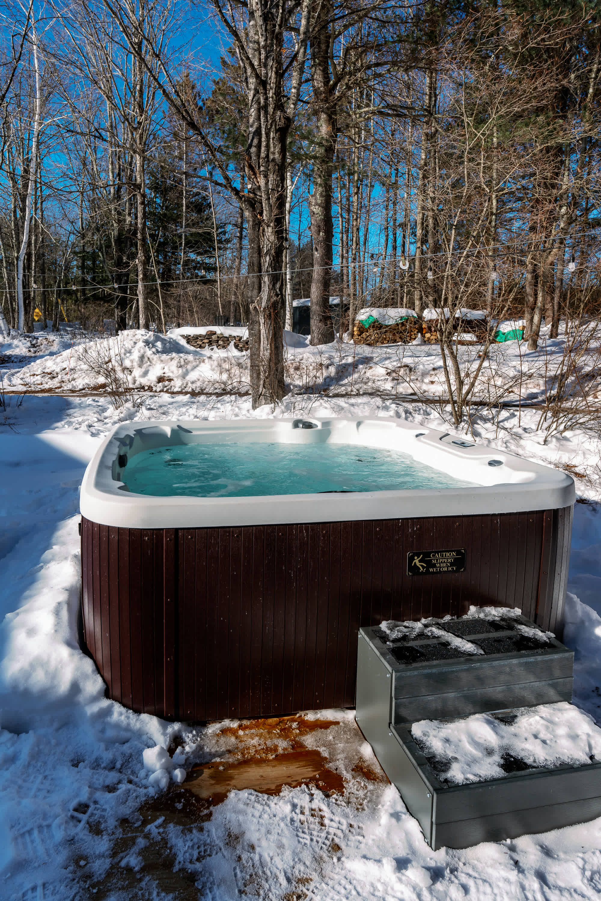 A dark brown hot tub with bubbling light blue water is set in a snow-covered yard, surrounded by bare trees under a clear blue sky. A dark grey step with snow on it leads up to the tub.