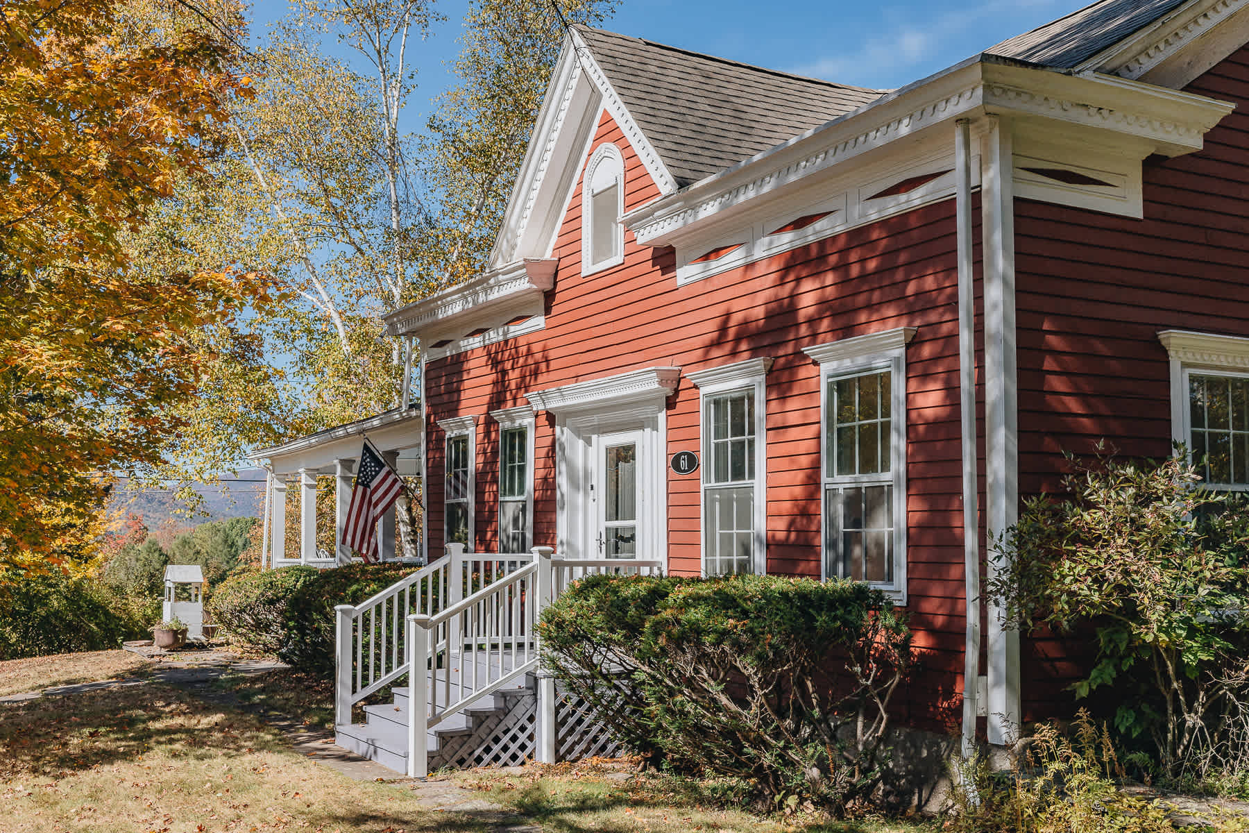 A charming red farmhouse with white trim and a gabled roof, featuring a welcoming front porch with white railings and an American flag. The house is surrounded by vibrant autumn foliage under a clear blue sky.