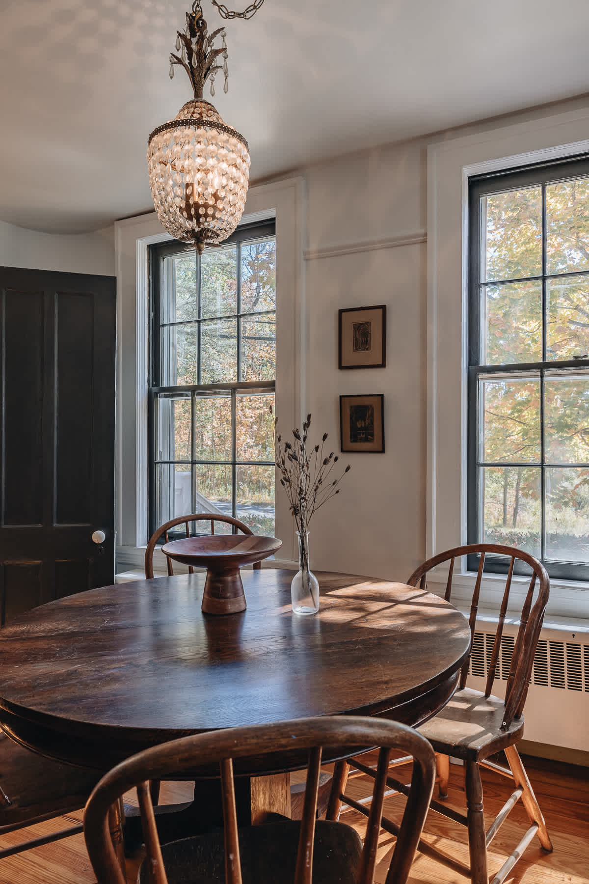 Sunny dining nook with a round dark wood table, matching wooden chairs, and an ornate crystal chandelier. Two large windows offer views of the outdoors.