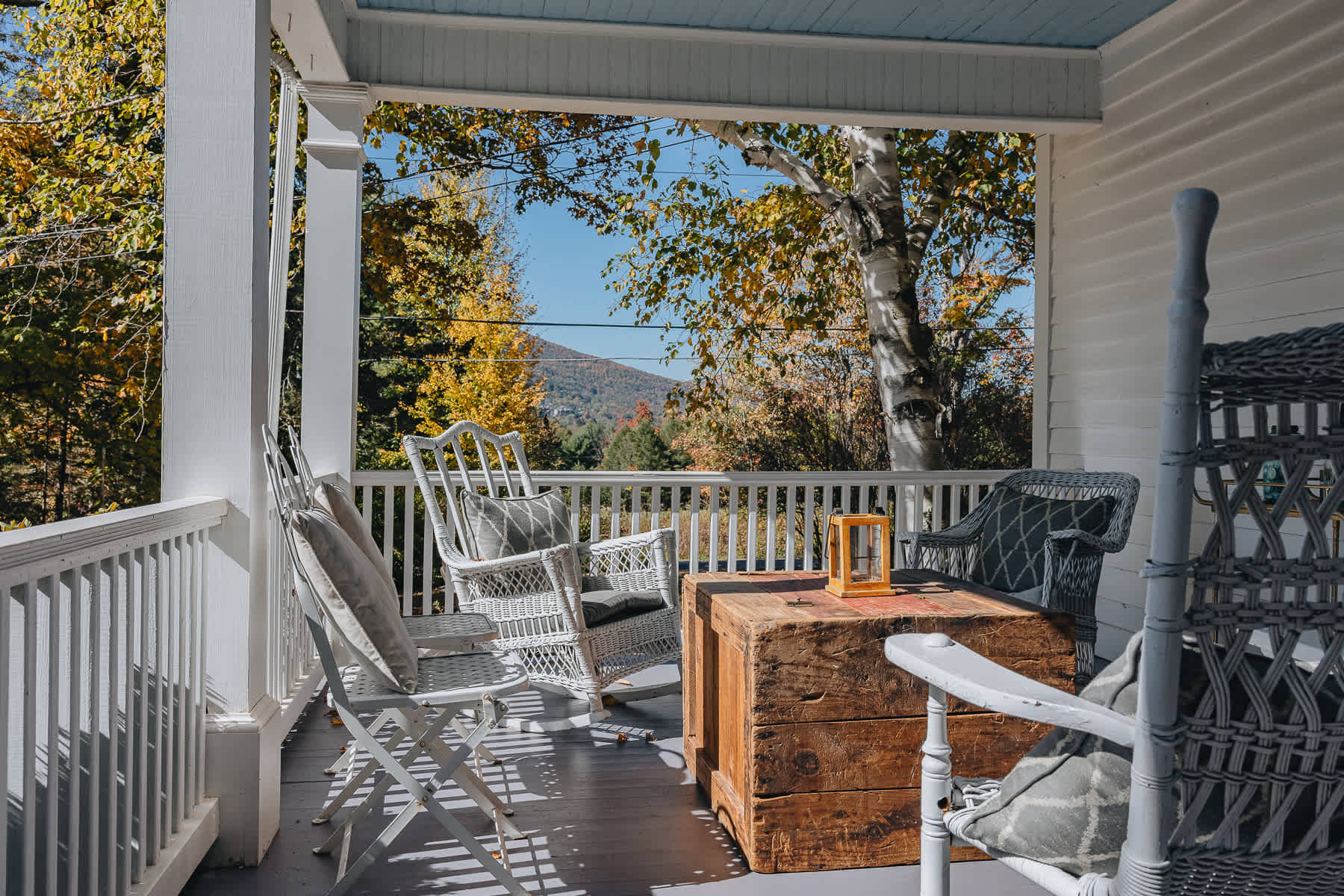 A cozy white porch furnished with wicker chairs and a wooden trunk table, offering a scenic view of distant mountains and colorful autumn trees. The porch ceiling is painted light blue.
