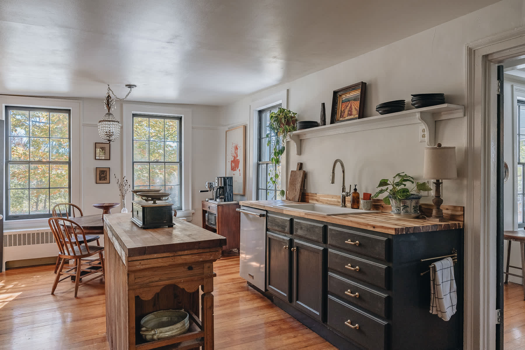 Bright kitchen with dark lower cabinets, butcher block countertops, a white farmhouse sink, and a rustic wooden island. Large windows overlook autumn foliage.