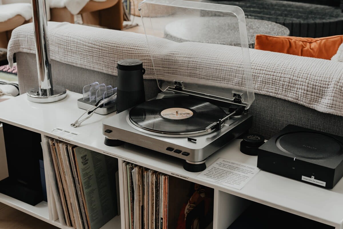 A modern entertainment setup features a silver turntable with a vinyl record playing, a black speaker, and a collection of vinyl records stored in the shelves below, all on a white console.