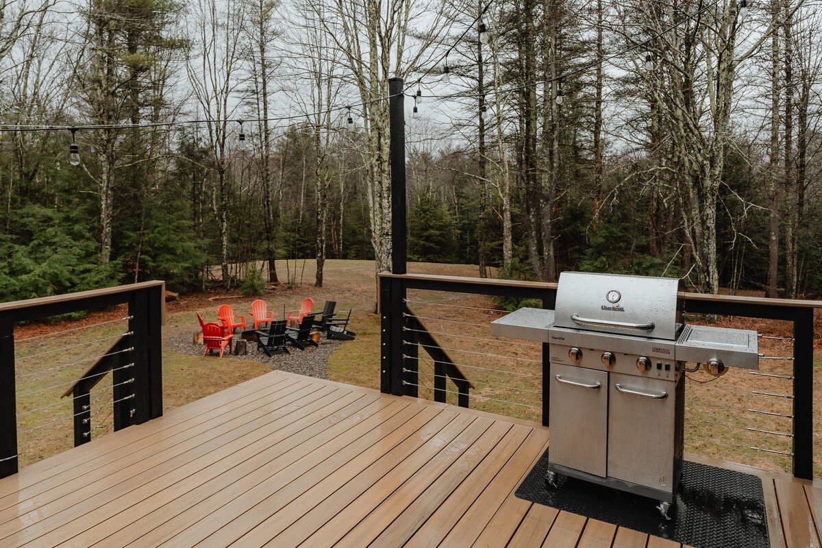 A spacious wooden deck features a stainless steel BBQ grill, overlooking a grassy yard with a gravel fire pit area surrounded by red and black Adirondack chairs, all backed by a dense forest.