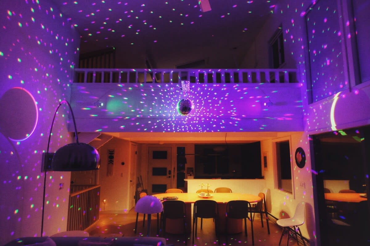 A vibrant game room with a disco ball casts colorful light patterns across the walls and ceiling. The two-story space features a dining area below and a lofted walkway above, creating a lively atmosphere.