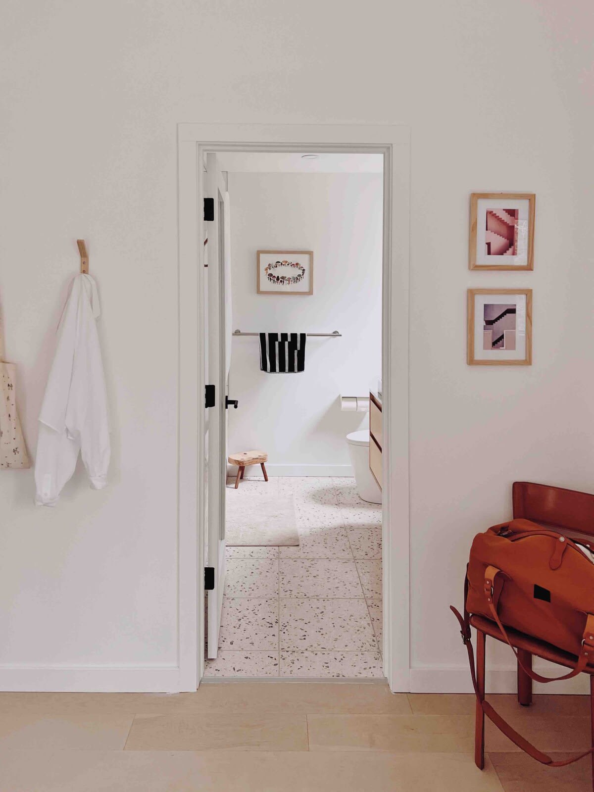A white doorway opens into a bathroom with speckled terrazzo flooring, a towel rack with a striped towel, and framed artwork, hinting at a toilet and vanity further inside.