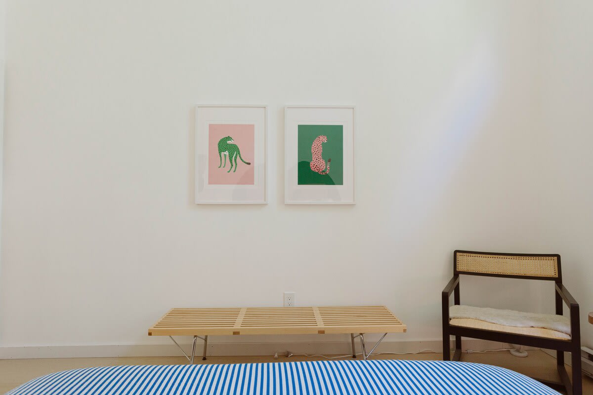 A minimalist bedroom with white walls, featuring two framed art prints of green leopards, a light wood slatted bench, and a dark wood chair with a cane back. A blue and white striped bed is partially visible.