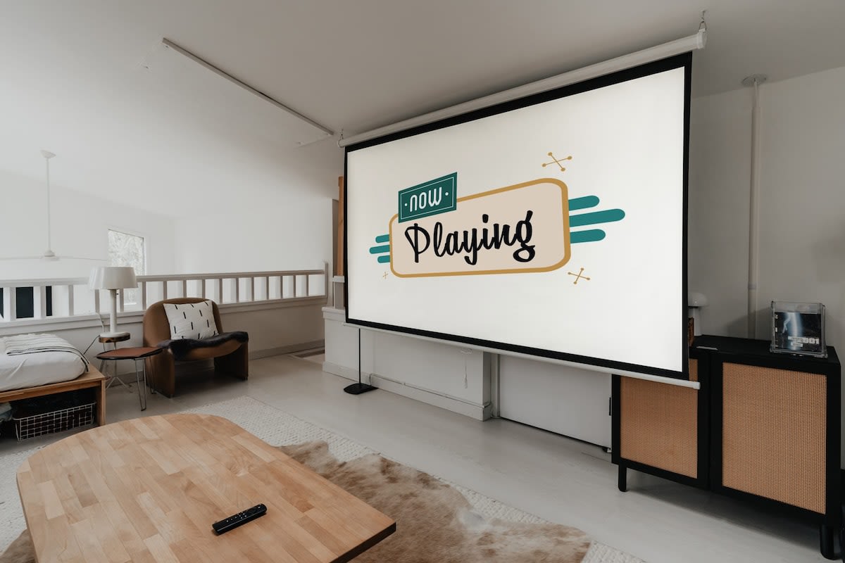 A bright media room features a large pull-down projector screen displaying a 'Now Playing' graphic. The room has light wood floors, a cowhide rug, and a modern wooden coffee table, with a lofted area visible in the background.