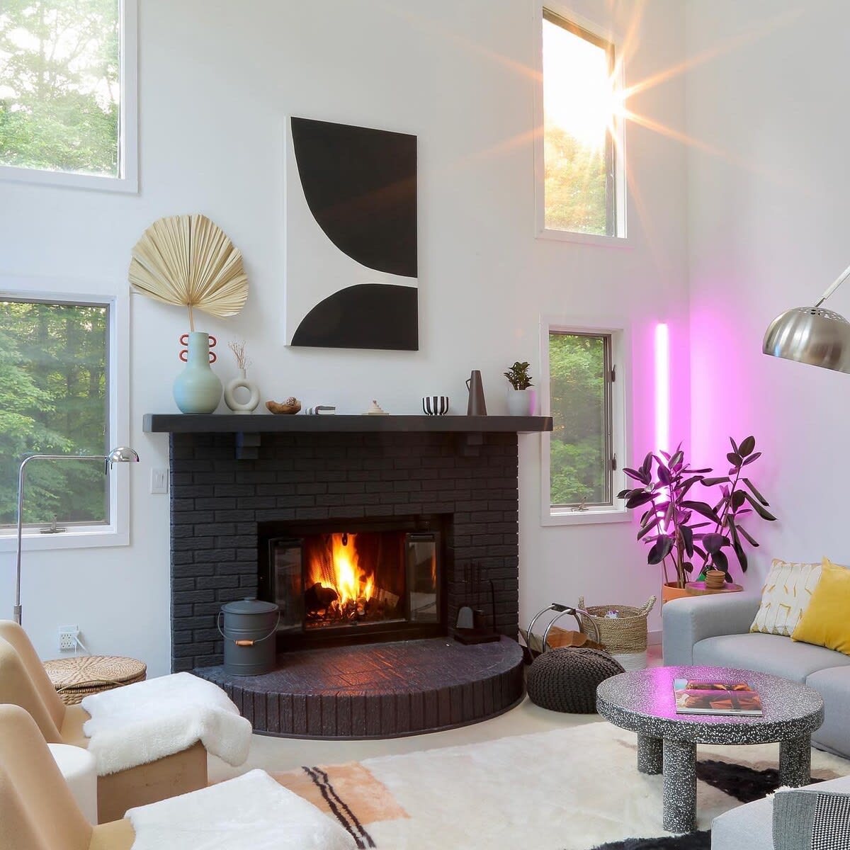 A bright and airy living room is centered around a black brick fireplace with a roaring fire. Modern abstract art hangs above, complemented by large windows, contemporary furniture, and a vibrant pink LED light strip.