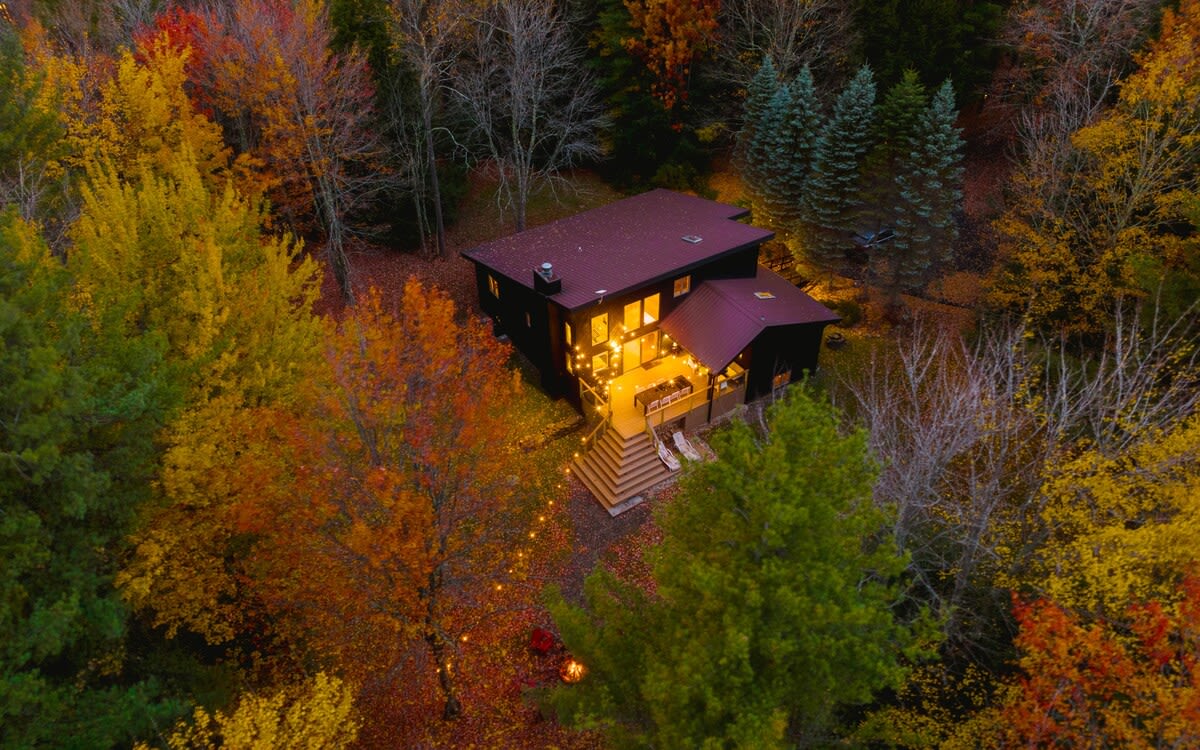 Aerial view of a dark modern house with a reddish-brown roof, nestled amidst a vibrant forest of red, orange, and yellow autumn trees. The illuminated deck and string lights are visible, along with a path leading to the house.