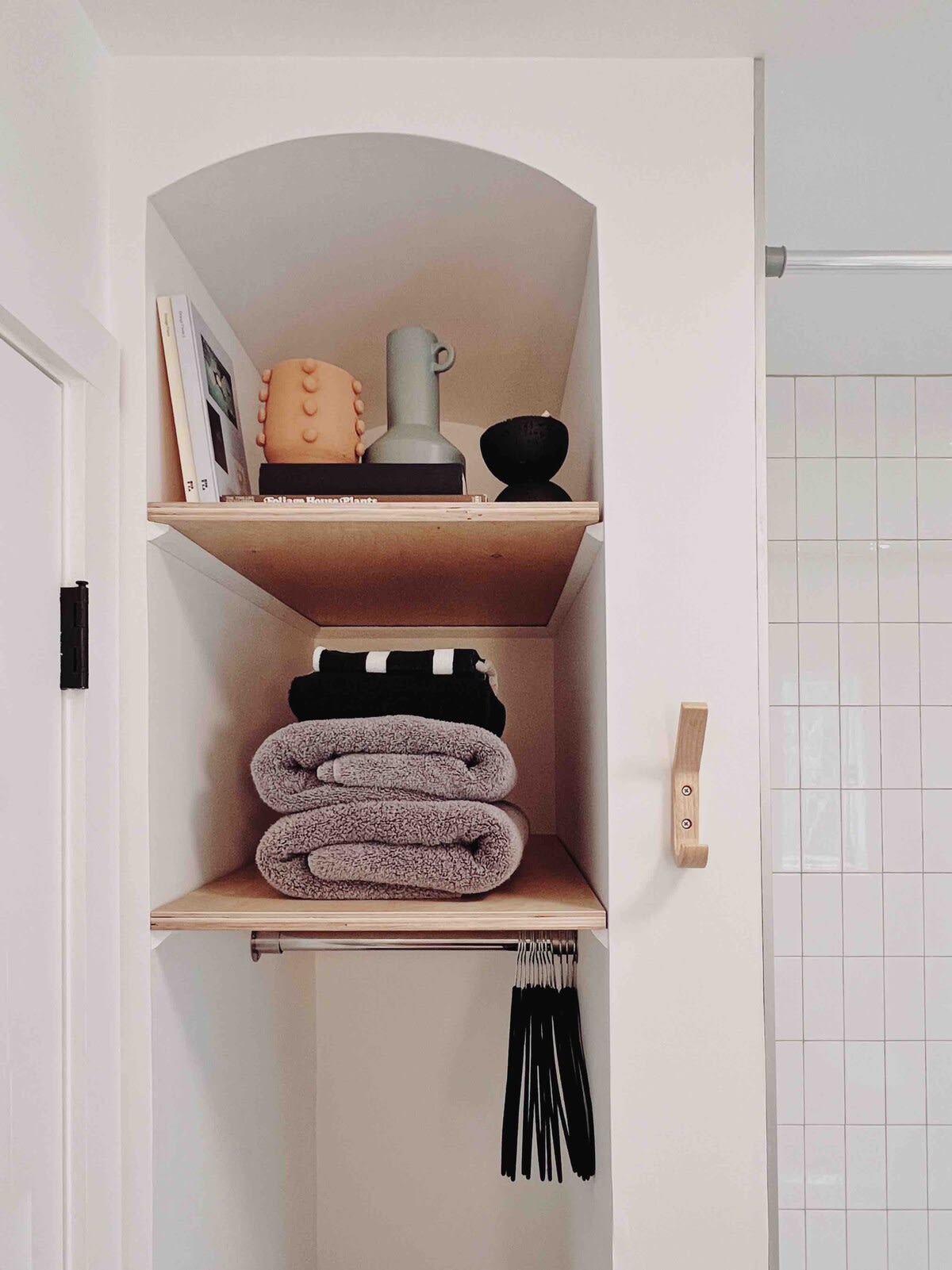 A built-in arched niche provides open shelving for decorative items and neatly folded towels, with a hanging rod below, set against white walls and light wood accents.