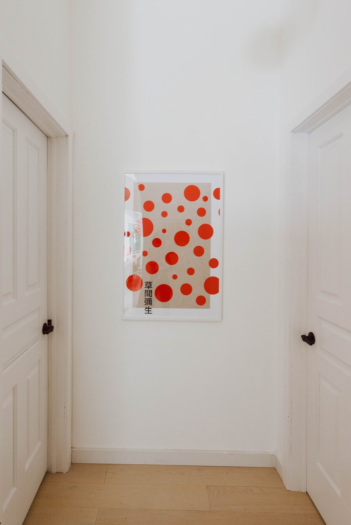 A minimalist white hallway features a framed modern art print with red polka dots and Japanese characters, flanked by two white doors with black handles.