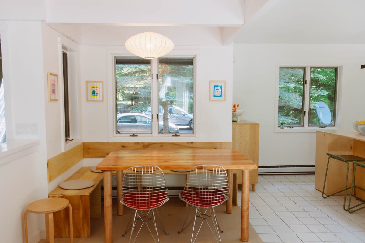A bright dining area features a wooden table with modern chairs, a built-in wooden bench, and a unique pendant light fixture. Large windows offer views of the outside, connecting to the adjacent kitchen area.