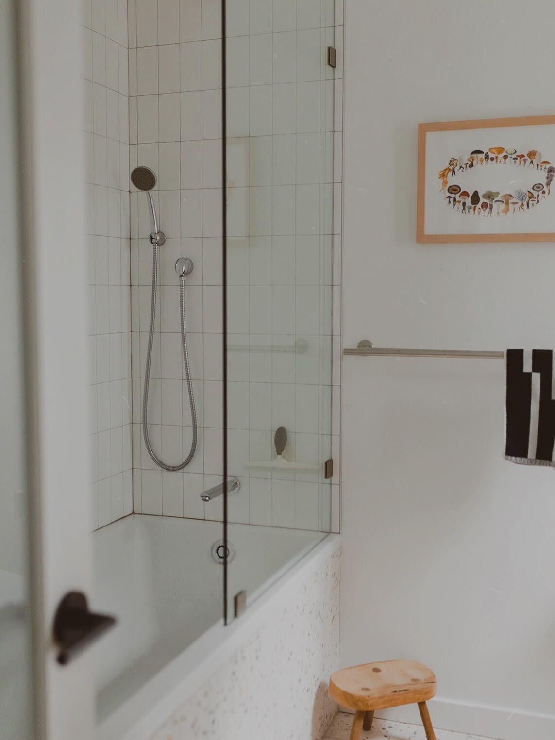 A clean bathroom showcases a white bathtub with a glass shower screen and a handheld showerhead, complemented by white subway tiles and a small wooden stool, with framed mushroom art on the wall.