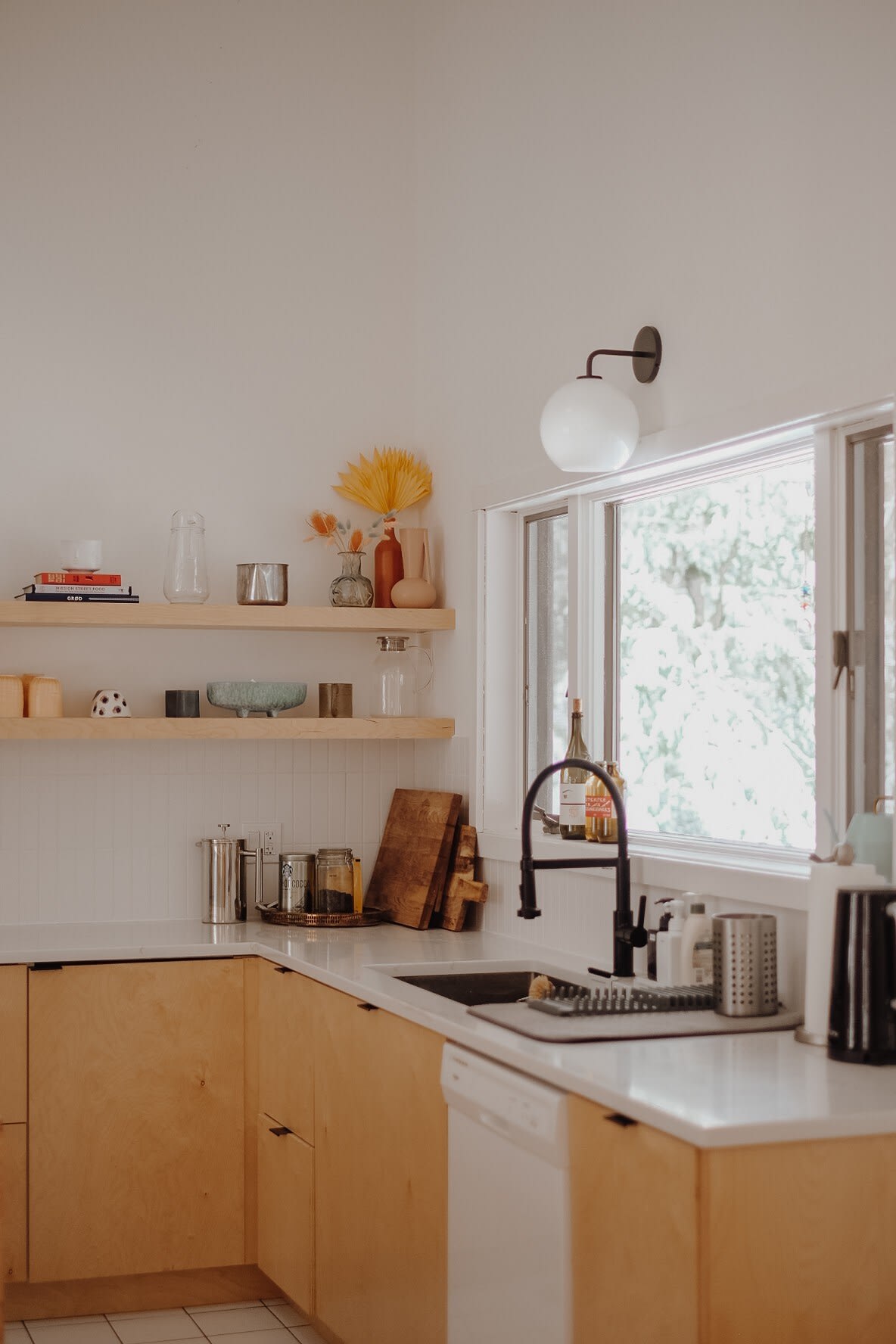 A bright corner of the kitchen features light wood lower cabinets, white countertops, and open shelving with various kitchen items. A large window overlooks green foliage, and a black faucet is installed over a sink.