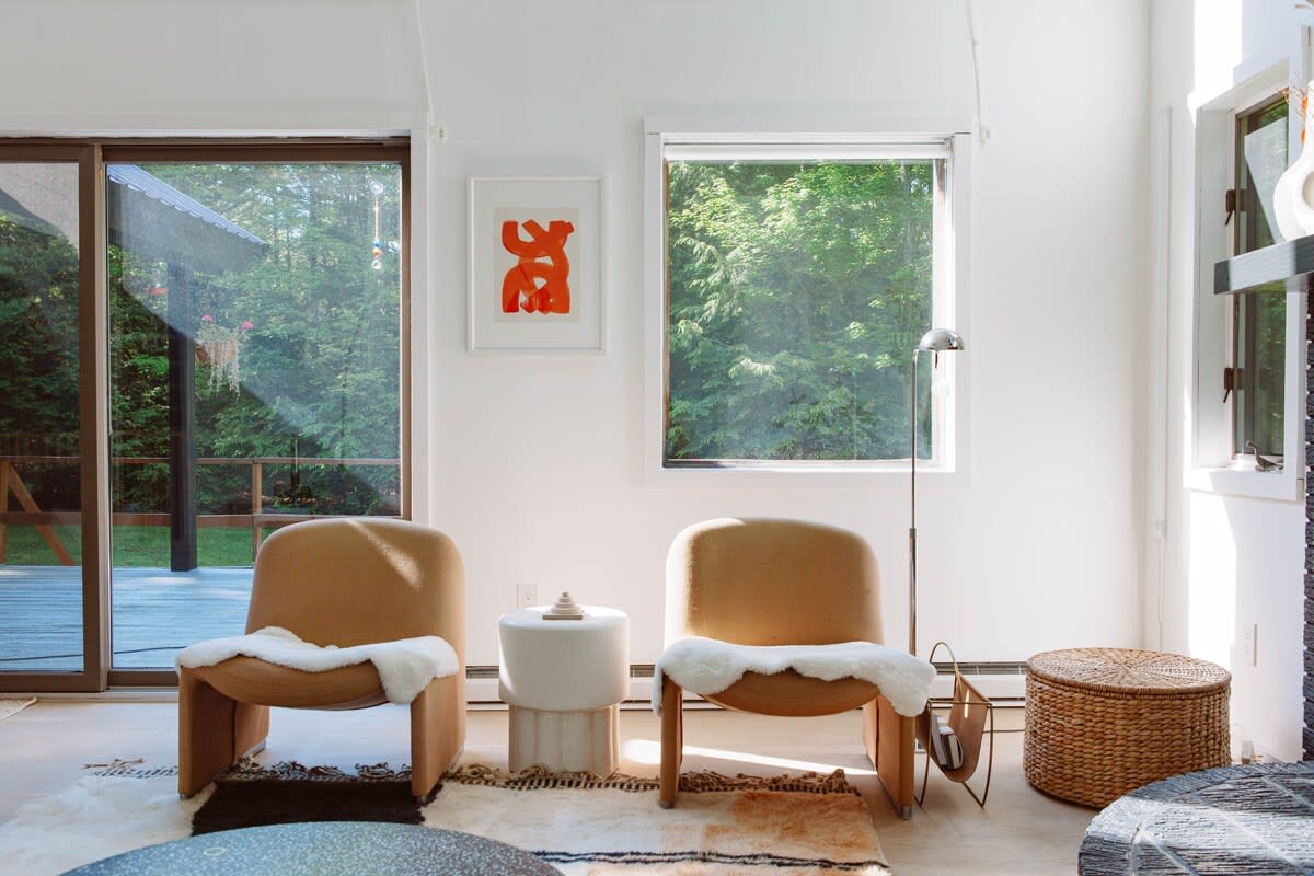 A bright living room features two modern tan armchairs with white sheepskin throws, a white side table, and a woven basket. Large sliding glass doors and a window offer views of lush green trees outside.