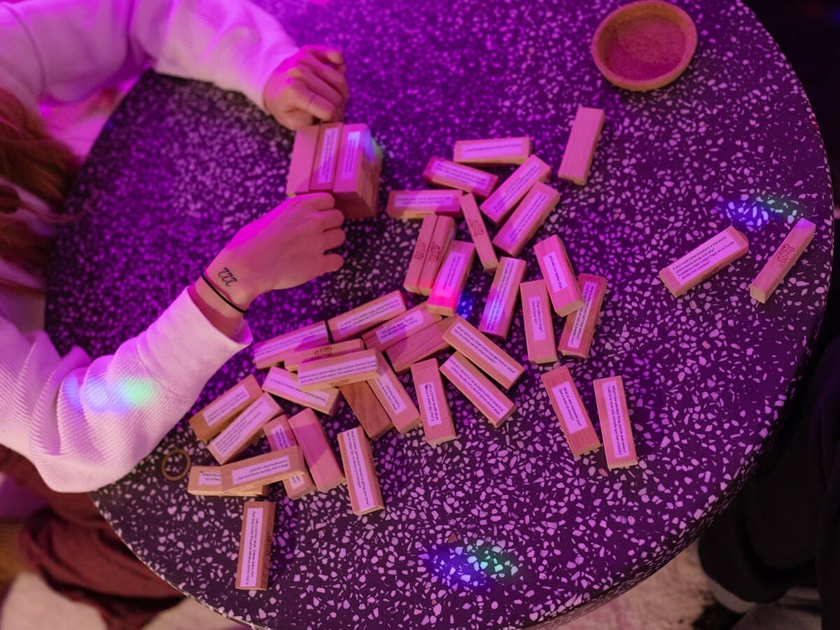 A person's hands are engaged in a board game with wooden blocks scattered on a dark, speckled table, illuminated by purple and pink ambient lighting.