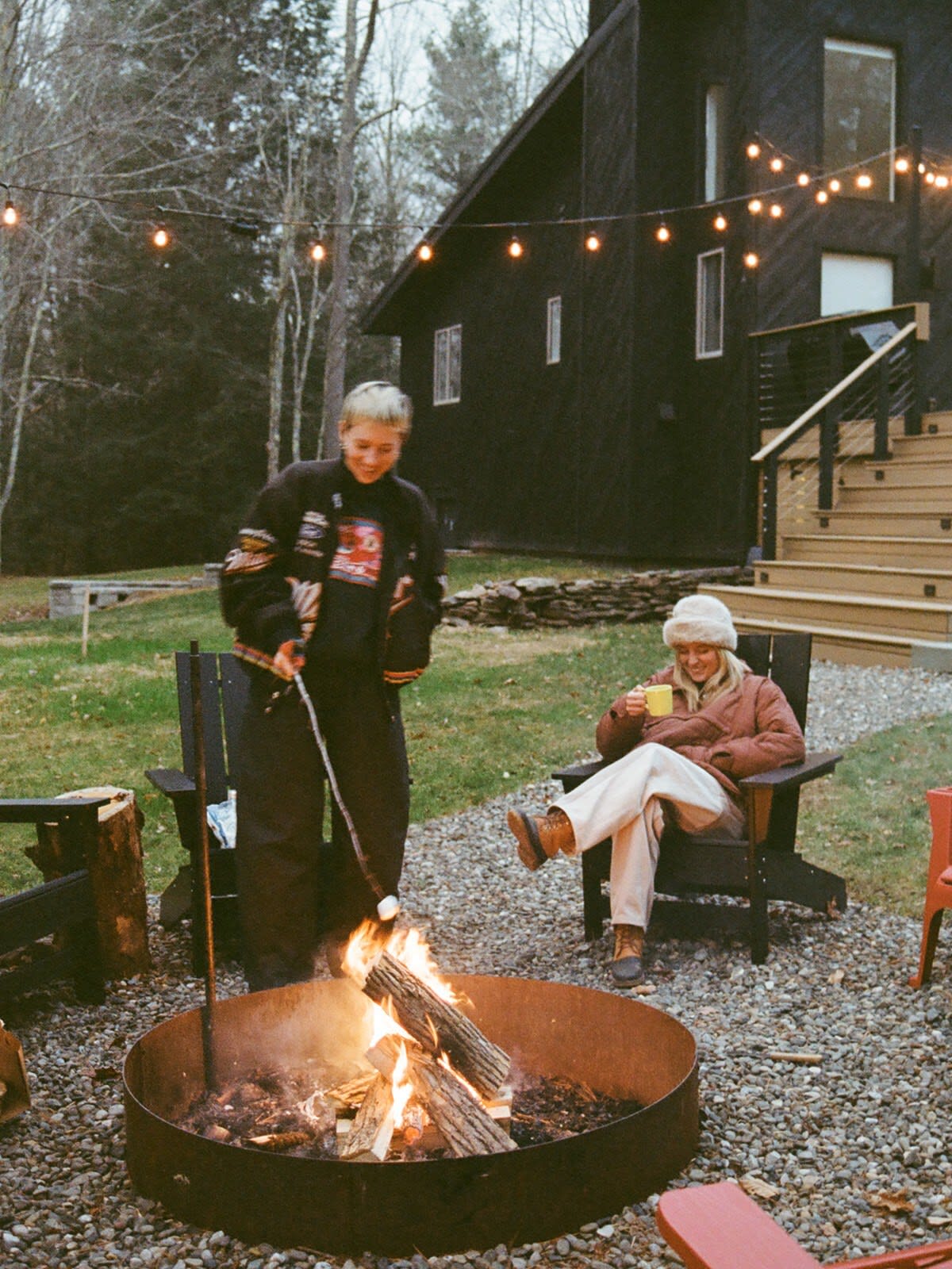 Two people are gathered around a metal fire pit with burning logs, roasting marshmallows in a gravel yard. String lights illuminate the scene, with a dark modern house visible in the background.