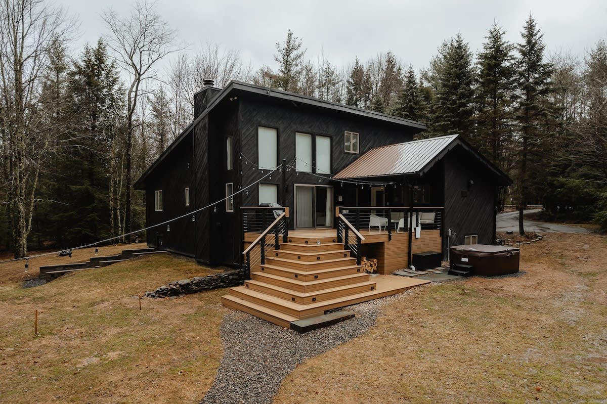 Modern black house with a spacious multi-level wooden deck, outdoor seating, and a hot tub visible on the lower right. The surrounding landscape features sparse trees and a gravel path leading to the deck.