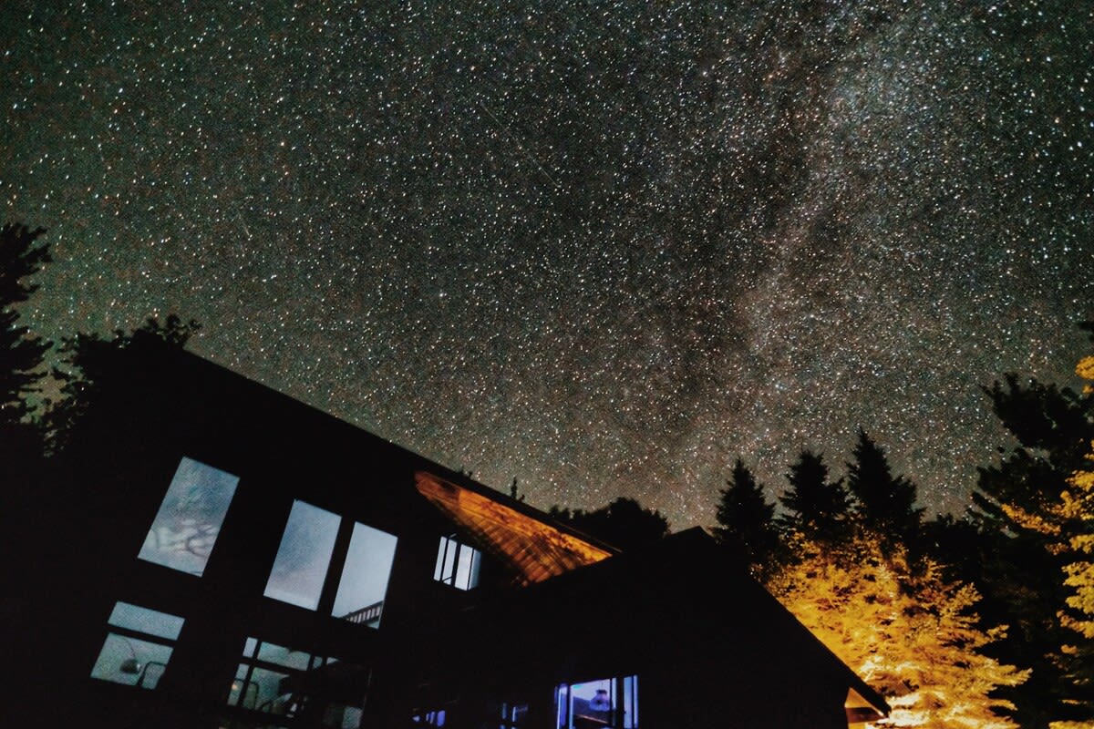Dramatic night shot of the dark house silhouetted against a brilliant starry sky, revealing the Milky Way. Interior lights glow from some windows, and tall trees frame the scene.