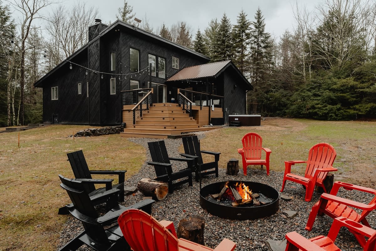 The exterior of a modern black cabin features multiple windows and a large wooden deck with stairs leading down to a fire pit area with red and black Adirondack chairs, surrounded by a forest.