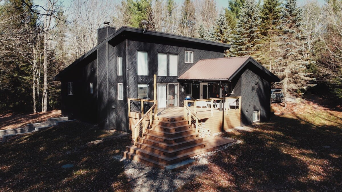 Daytime view of the modern black house with a multi-level wooden deck, featuring outdoor seating and a grill. The surrounding bare trees suggest late autumn or early spring, with some evergreen trees in the background.