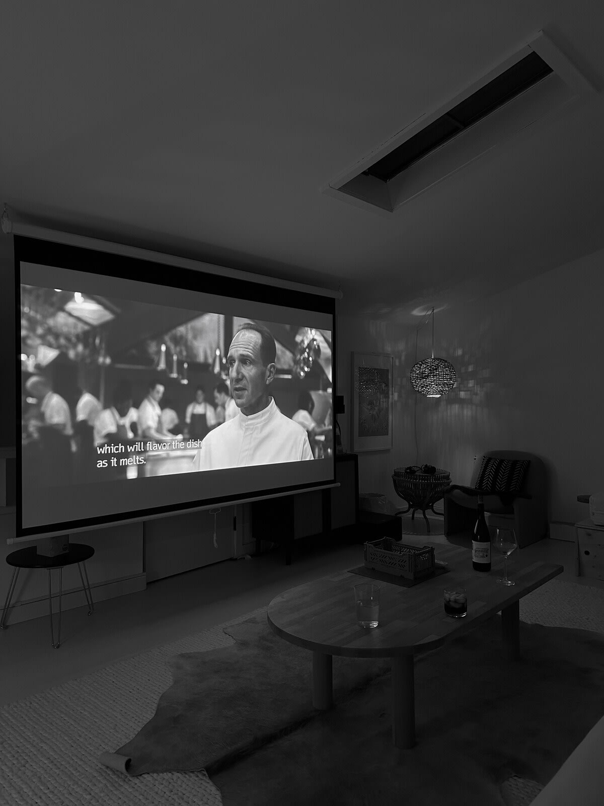 A darkened media room shows a large projection screen displaying a movie scene with subtitles, with a wooden coffee table and a cowhide rug visible in the foreground.