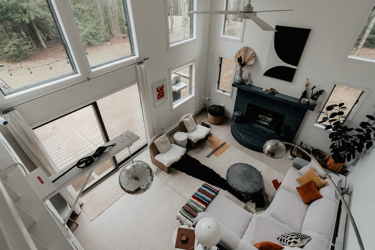 An aerial view of the bright living room shows a white sectional sofa, two armchairs, and a dark brick fireplace. A large disco ball hangs near the sliding glass doors, which open to a deck overlooking the forest.