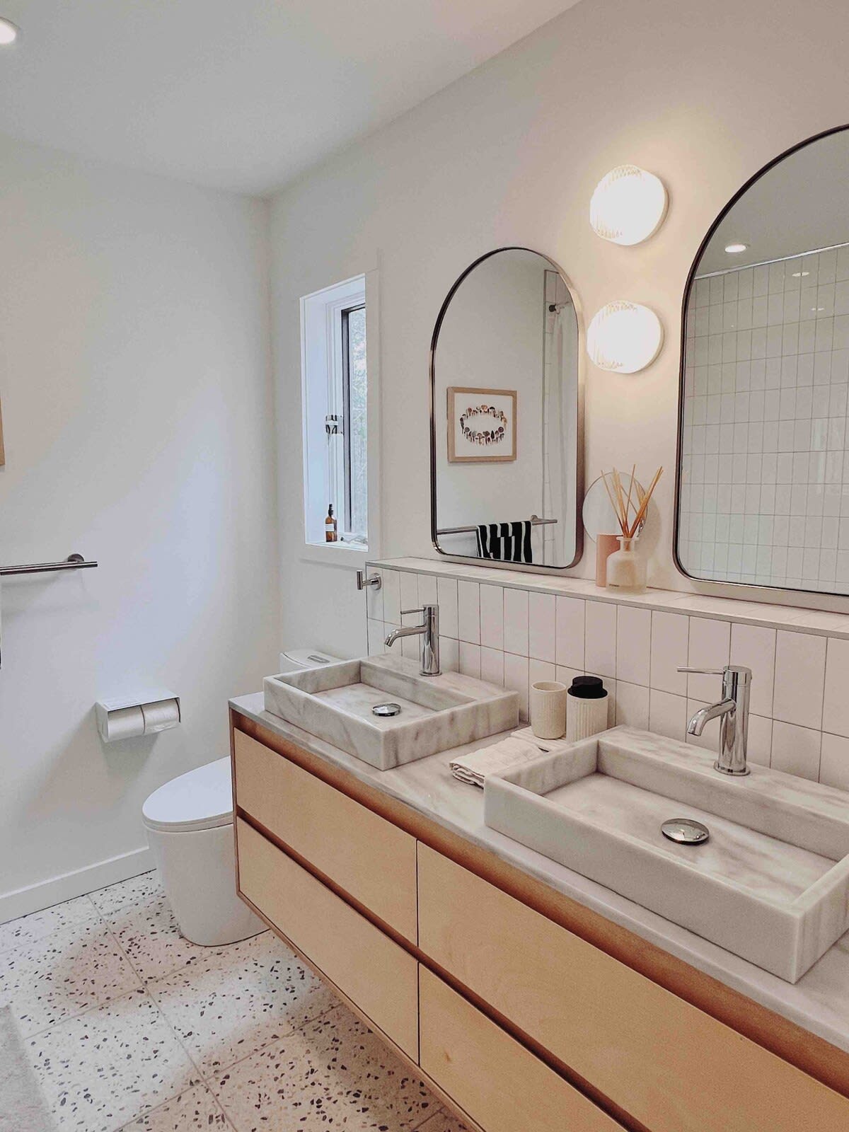 A modern bathroom features a double vanity with light wood cabinets and rectangular marble vessel sinks, flanked by arched mirrors and circular wall sconces, all set against white subway tile and terrazzo flooring.