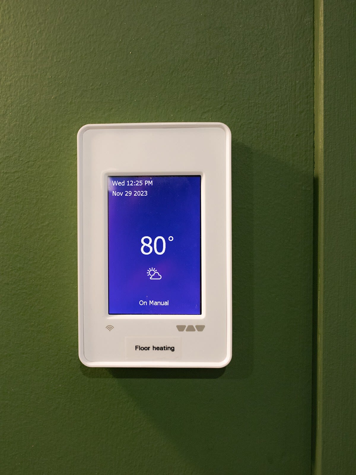 A close-up of a modern white digital thermostat displaying "80°" and "Floor heating" on a dark green wall, showing the date and time as "Wed 12:25 PM Nov 29 2023".