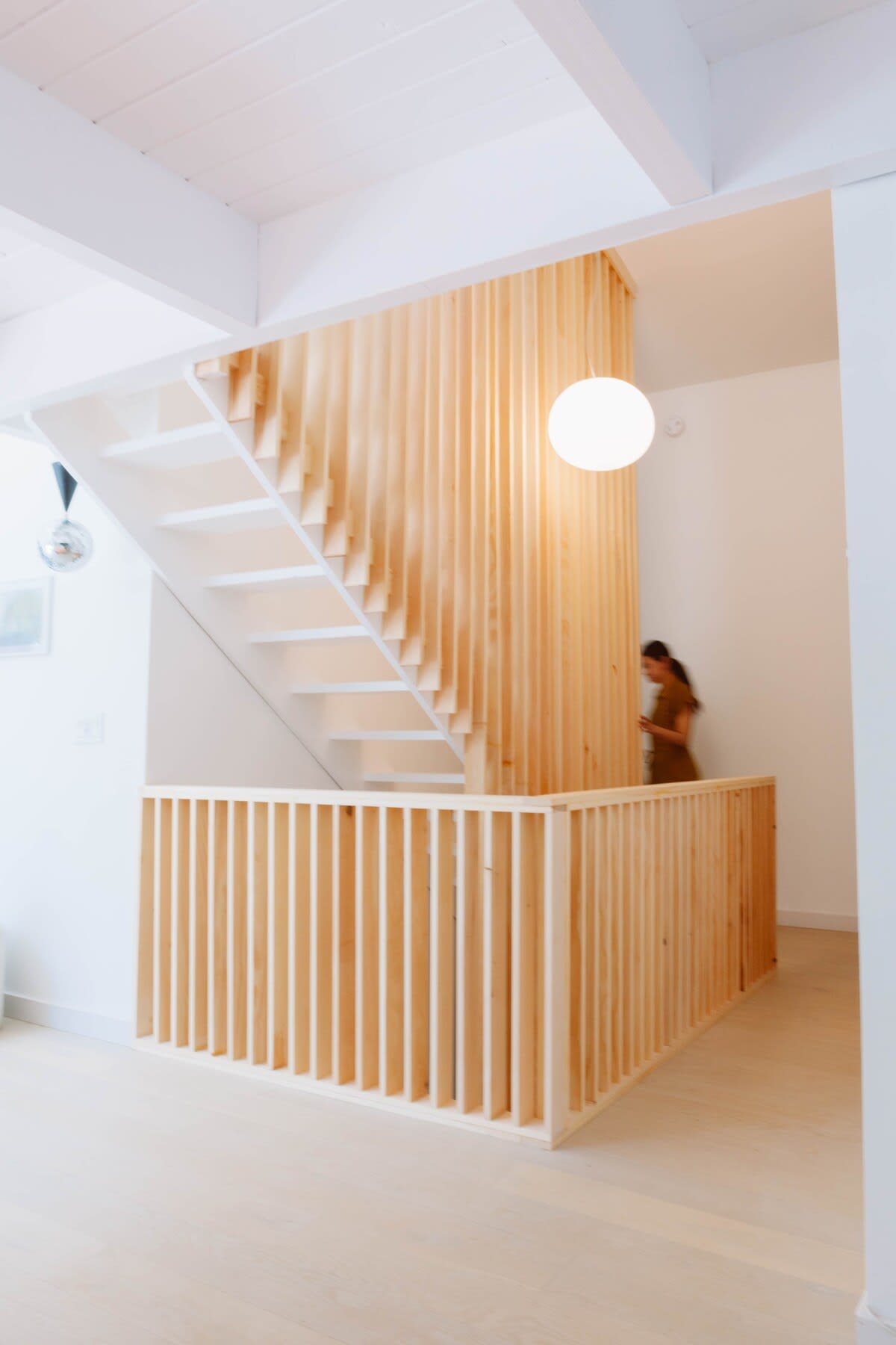 A modern, light-filled foyer showcases a striking white staircase with natural wood slat railings, extending from the ground floor to an upper level, illuminated by a spherical pendant light.