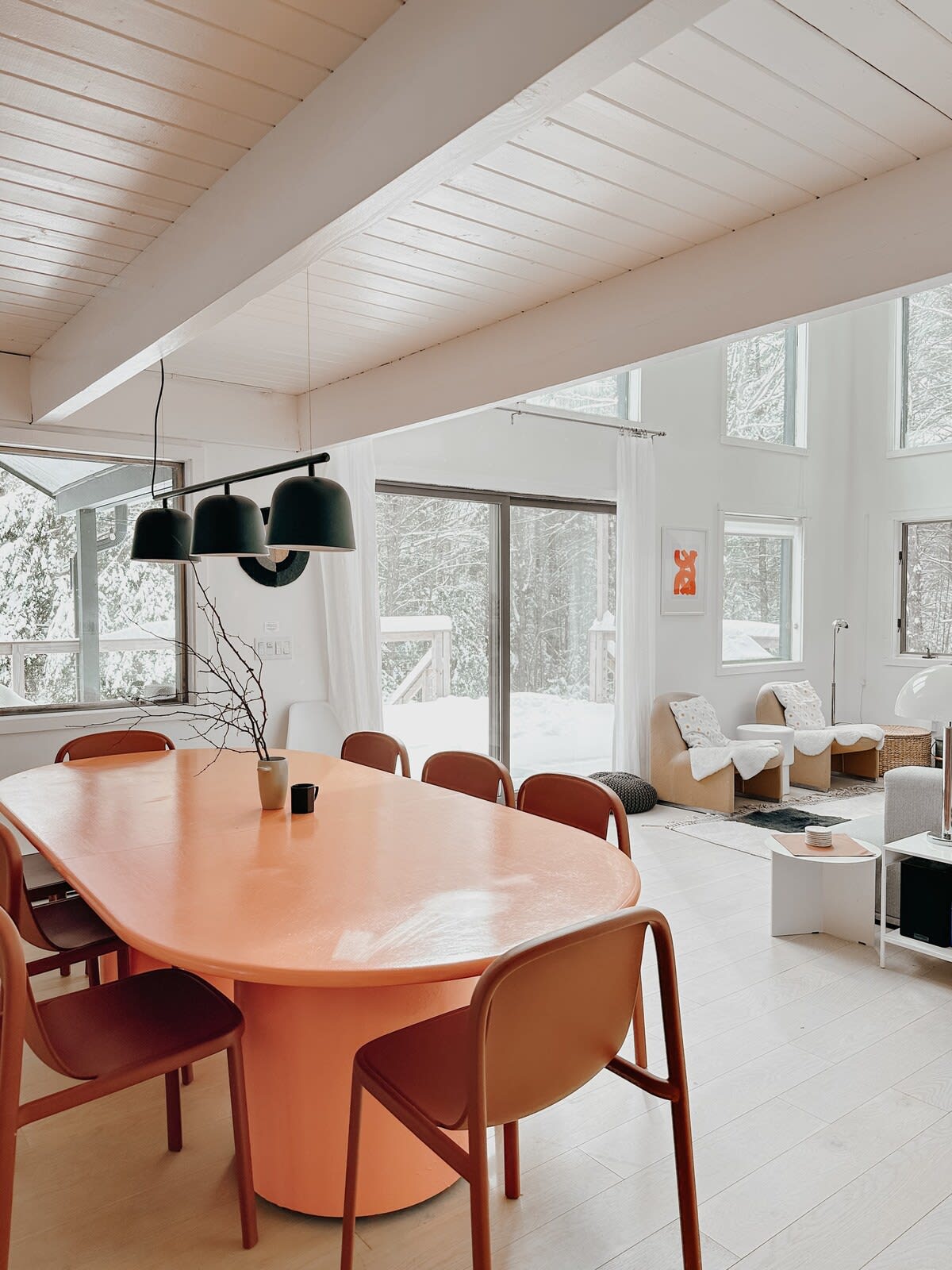 A bright, open-concept dining area features a large, vibrant orange oval table surrounded by matching rust-colored chairs, set under a modern black linear pendant light. The space opens to a living area with large windows overlooking a snowy landscape.