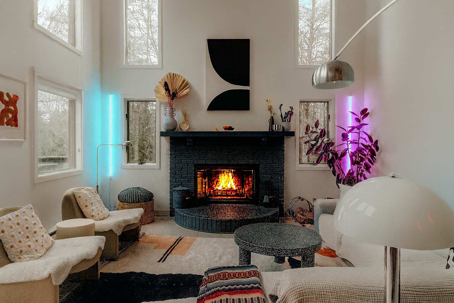 A bright living room features a black brick fireplace, modern abstract art, and large windows. Colorful LED strip lights add a vibrant glow, complementing the contemporary furniture and decor.