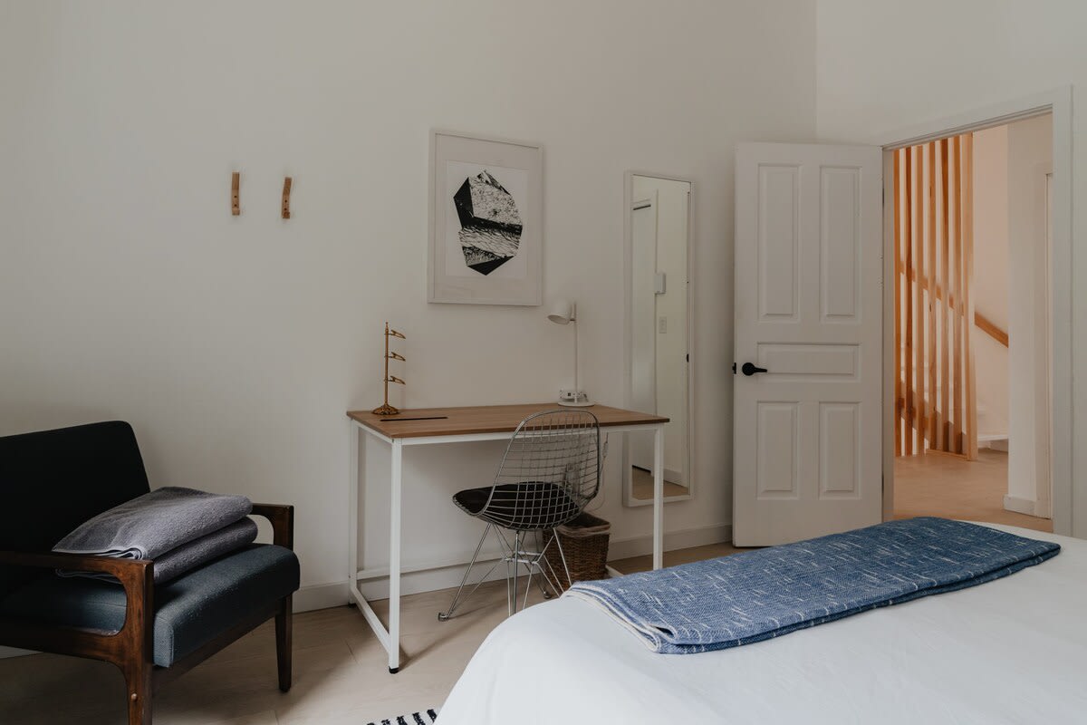 A bright bedroom corner featuring a light wood desk with a metal wire chair, a full-length mirror, and a white paneled door. The white walls are adorned with a framed abstract print and wooden wall hooks.