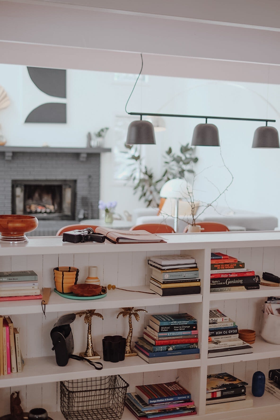 A white built-in bookshelf filled with books and decorative items acts as a room divider, offering a glimpse into an open living space with a gray brick fireplace and a modern black pendant light.