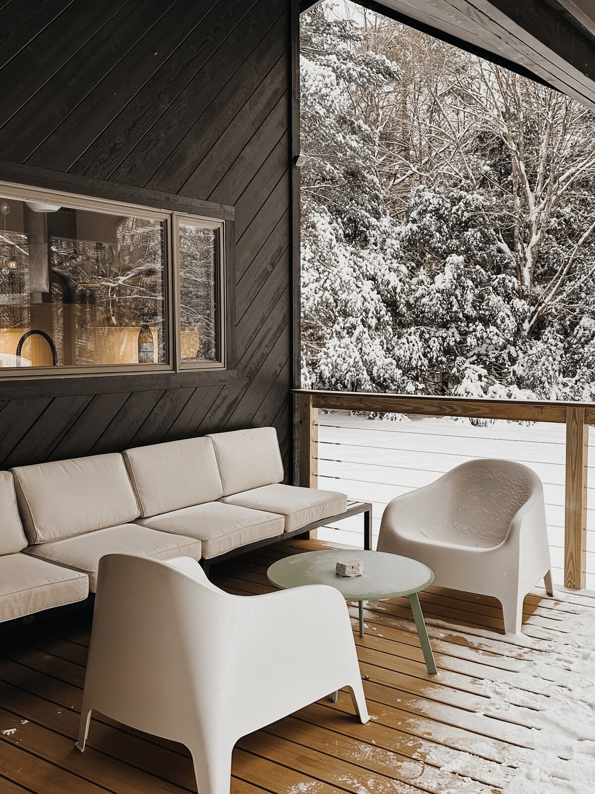 The outdoor deck is covered in a light dusting of snow, featuring a white sofa and armchairs around a light green coffee table, with a snowy forest visible beyond the railing.