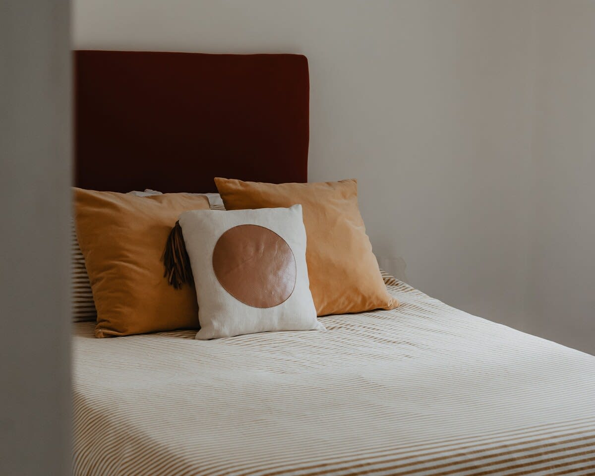 Close-up of a bed with a reddish-brown upholstered headboard, adorned with striped beige and white bedding, and decorative pillows in mustard yellow and a cream pillow with a brown leather circle.