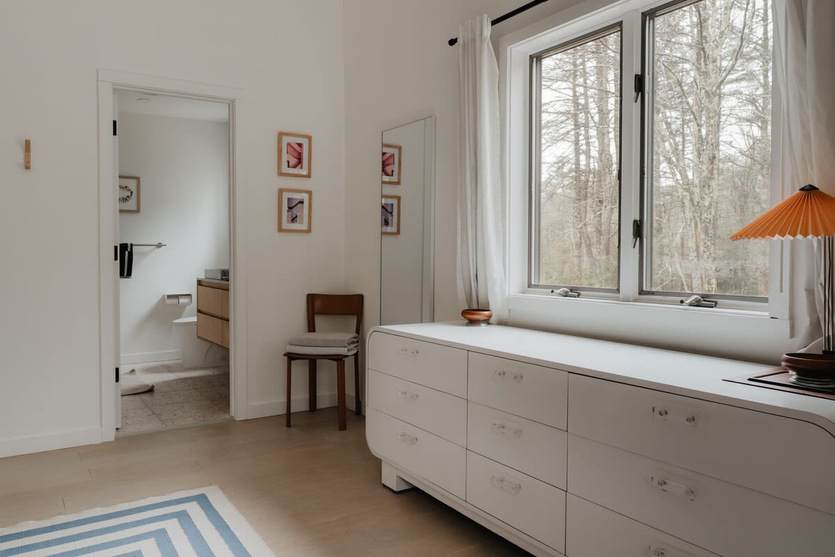A bright bedroom features a white dresser with multiple drawers, a full-length mirror, and framed art on the wall, with a doorway revealing a bathroom with a toilet and vanity.