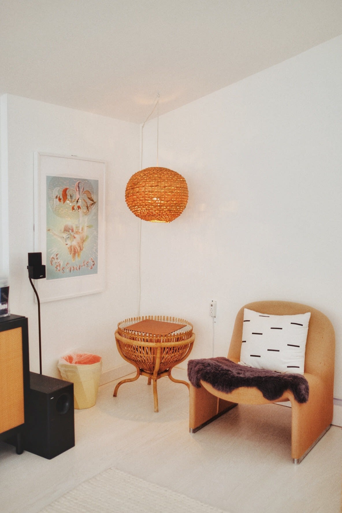 A bright corner of a living space features a modern, curved tan armchair with a dark faux fur throw, a woven spherical pendant light, and a small round wicker table.