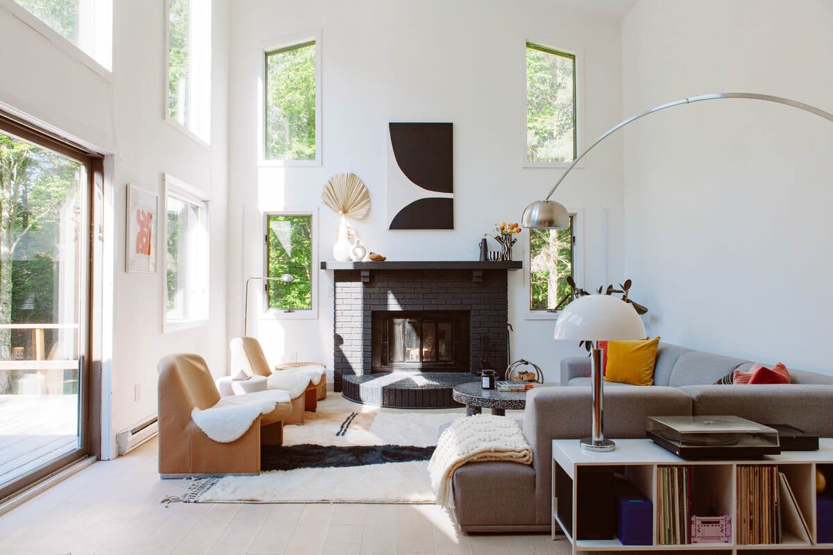 A spacious, sunlit living room features a dark brick fireplace, a grey sectional sofa, and two tan armchairs with white throws. Large windows and sliding glass doors provide ample natural light and views of the surrounding forest.