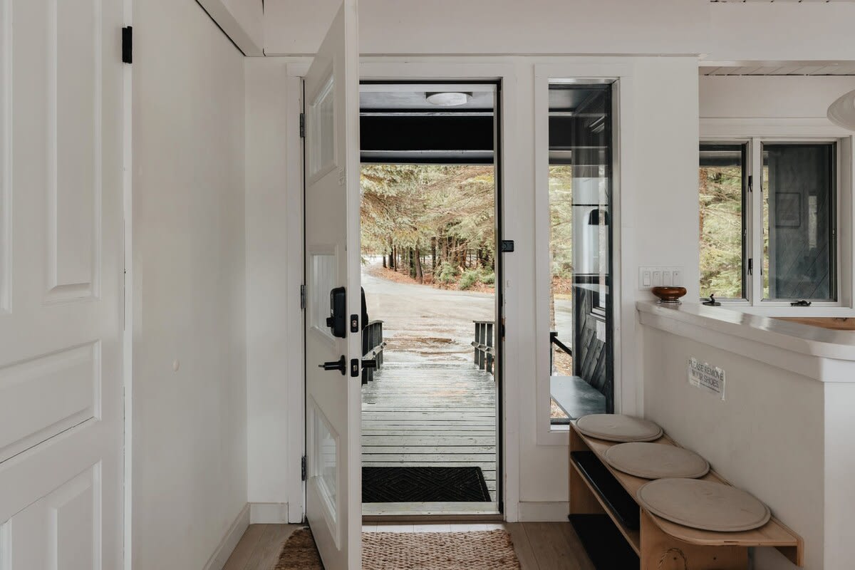 A bright and minimalist foyer features a white open door leading to an outdoor deck with views of a wooded area, a wooden bench with round cushions, and light wood flooring.
