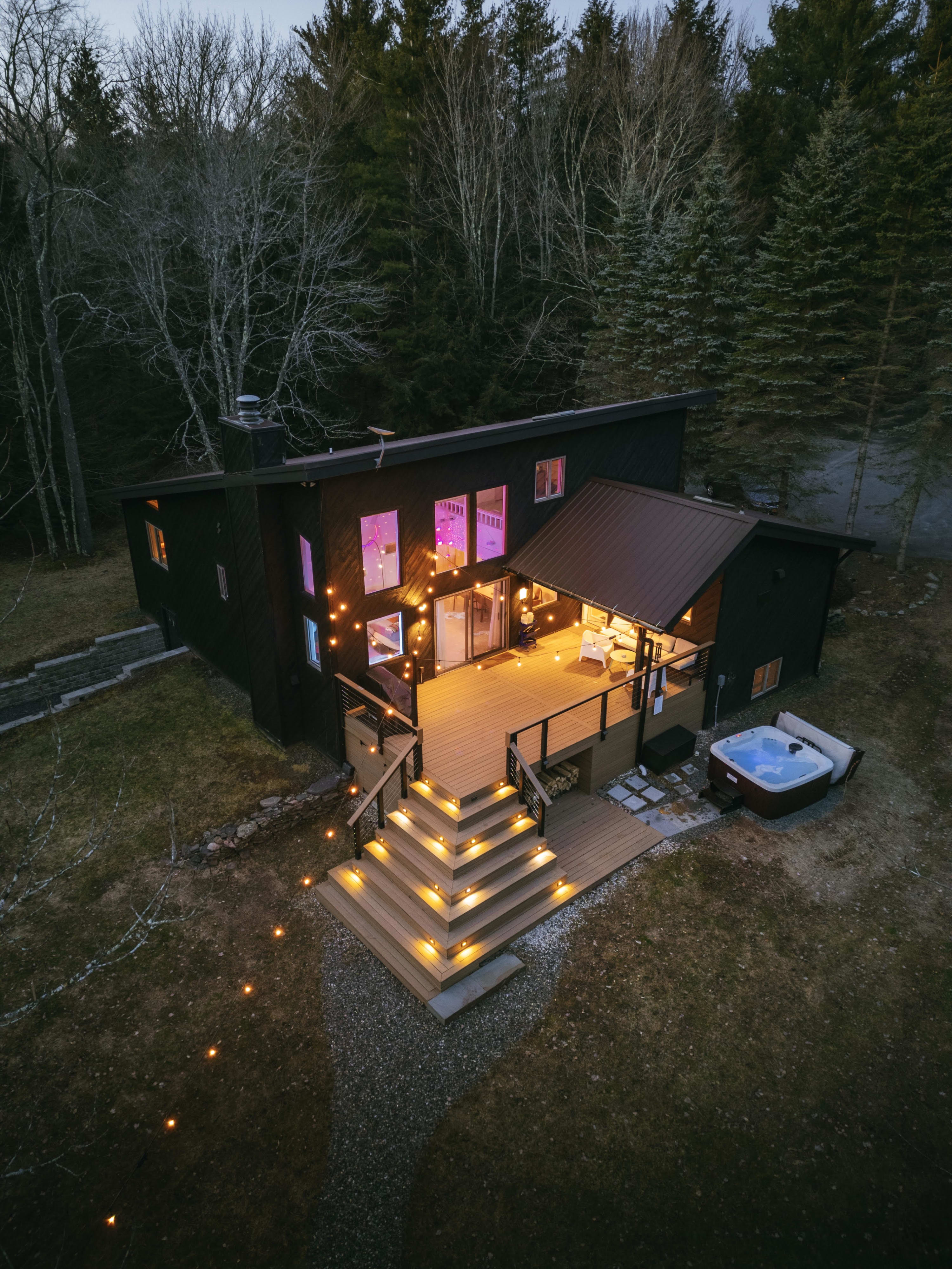 An aerial view of a modern black cabin nestled among evergreen and bare trees, featuring a large illuminated wooden deck and a hot tub. String lights adorn the deck and stairs, creating a warm glow against the twilight sky.
