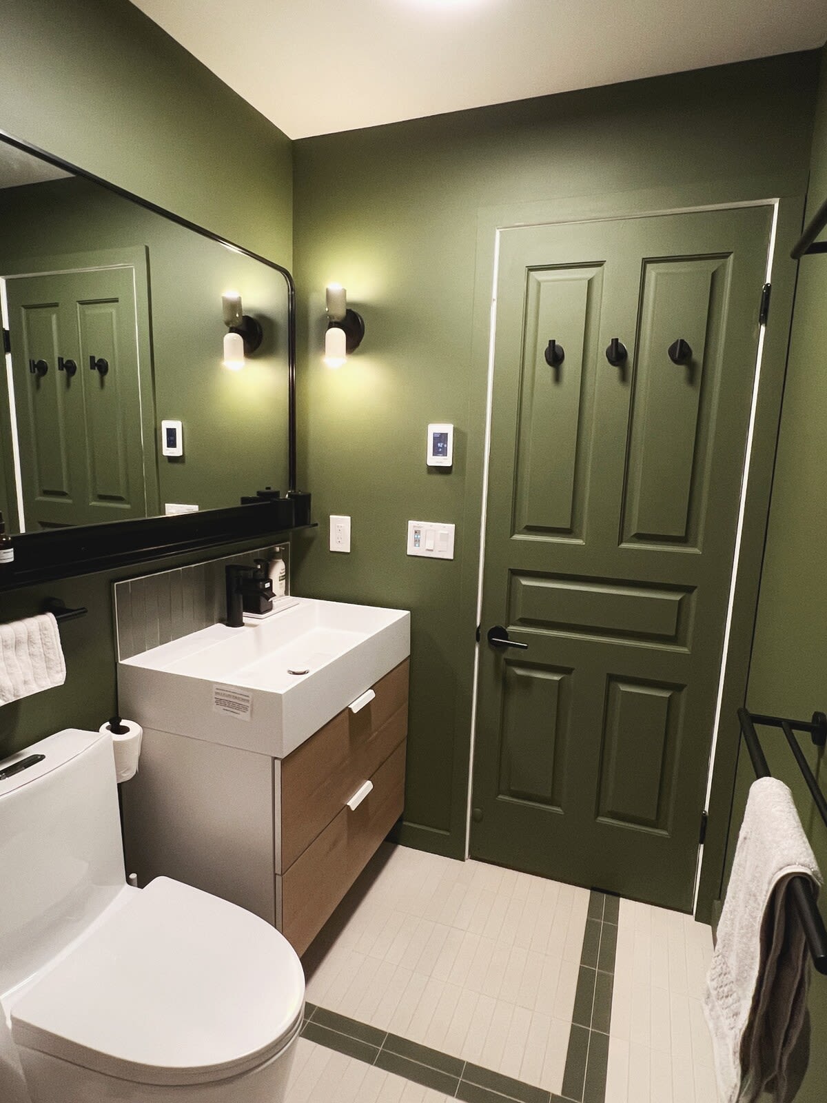 Contemporary bathroom with olive green walls, a white rectangular sink vanity with wooden drawers, and a large black-framed mirror. A white toilet and a matching green door with black hardware are also present.