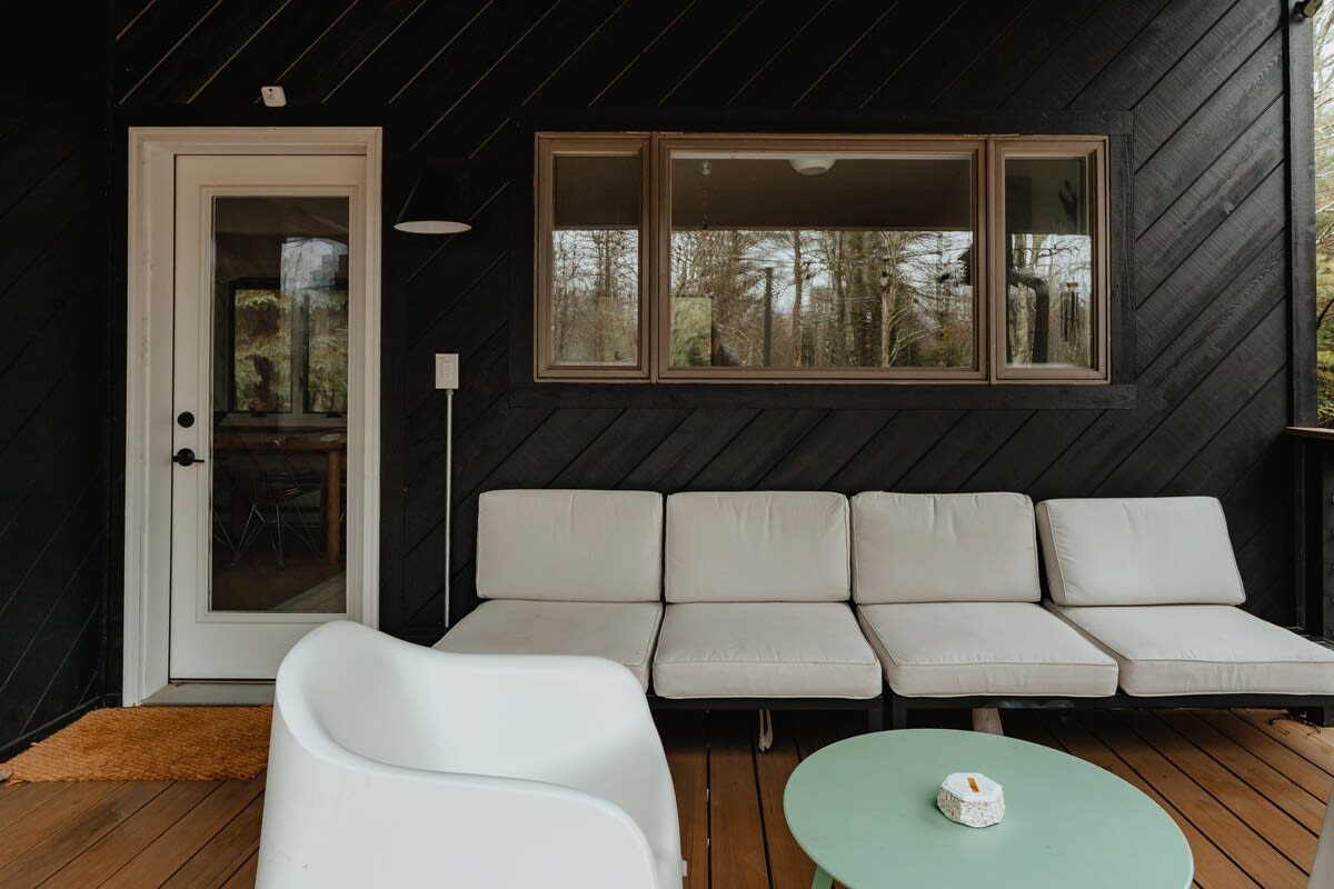 A covered outdoor deck features a long white sofa with cushions and a white armchair, set on a wooden floor against a dark wood-paneled wall, with a large window overlooking trees.