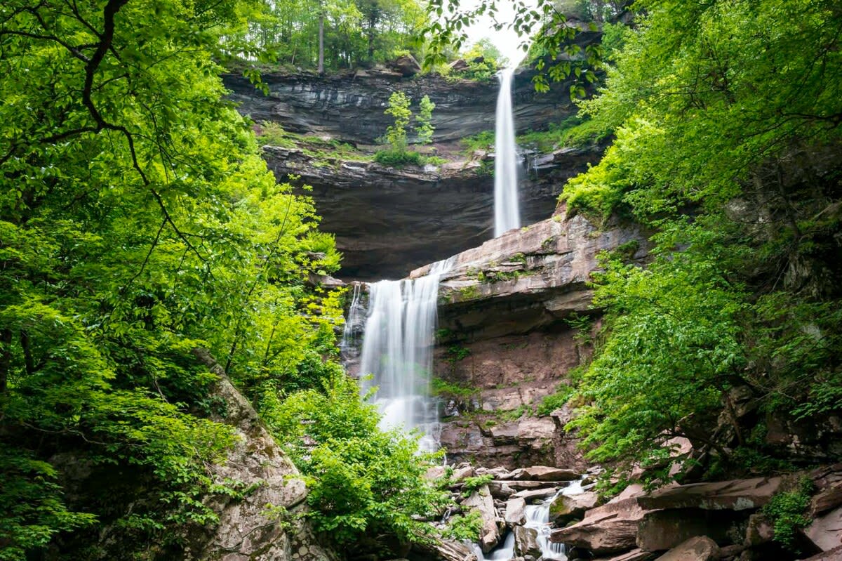A majestic two-tiered waterfall cascades down a rocky cliff face, surrounded by vibrant green deciduous trees and lush foliage under a bright sky.