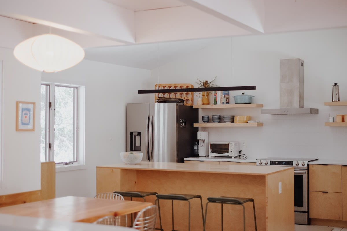 A modern kitchen with light wood cabinetry, open shelving, and stainless steel appliances. A large island with bar stools provides seating, illuminated by a unique pendant light.