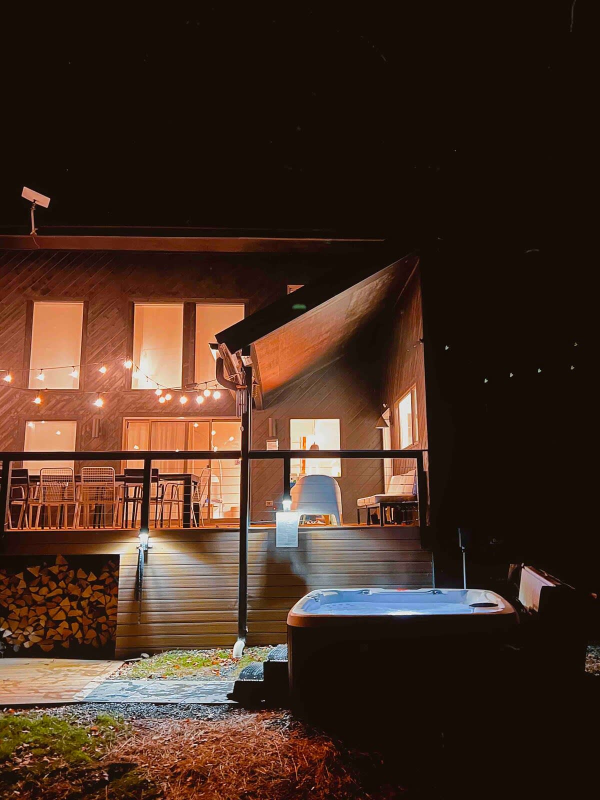 A hot tub is positioned on a wooden deck at night, with a stack of firewood nearby. The modern house exterior is illuminated by warm interior lights and string lights on the deck.
