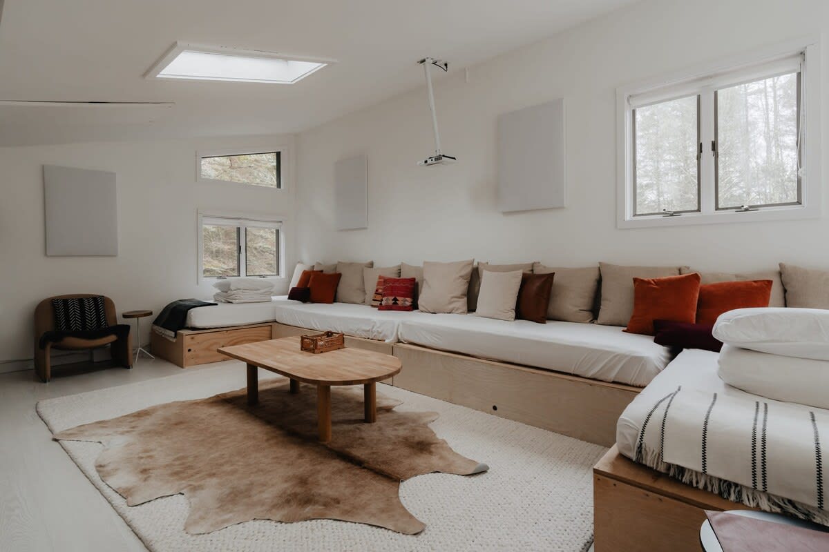 A cozy media room features built-in platform seating with numerous cushions in neutral and rust tones, a wooden coffee table, and a large cowhide rug over a textured area rug, with multiple windows.